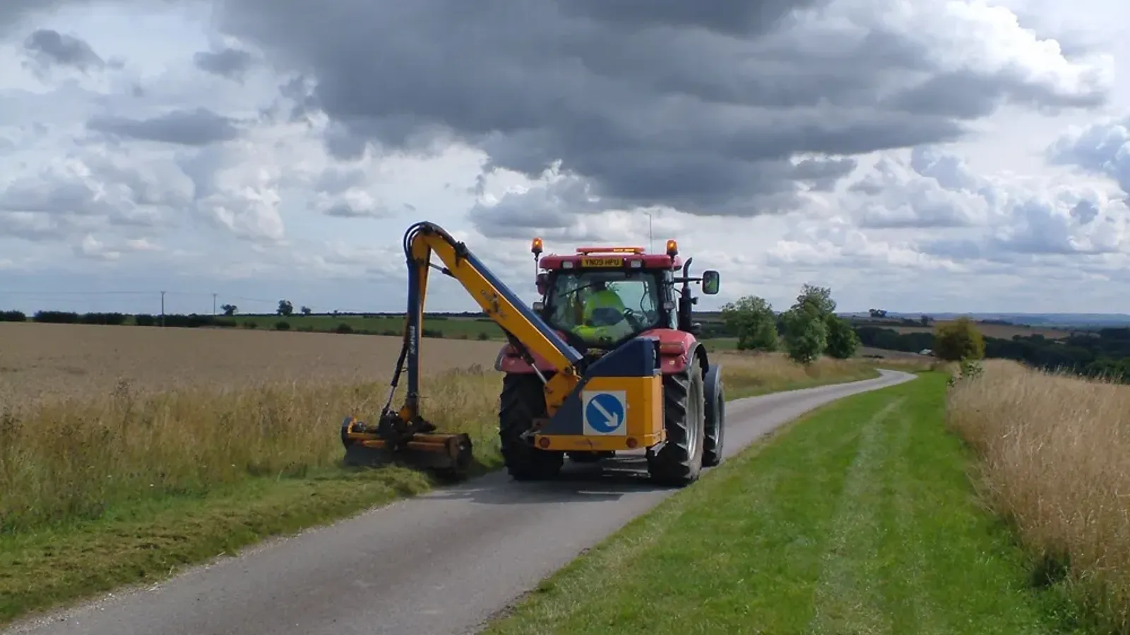 Tractor with a side-arm boom flail mower cutting roadside vegetation along a rural lane